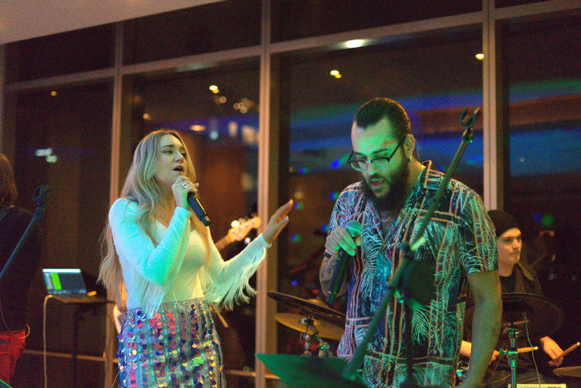 The full band across a window-lined room, keys and drums at the back, green light on the ceiling